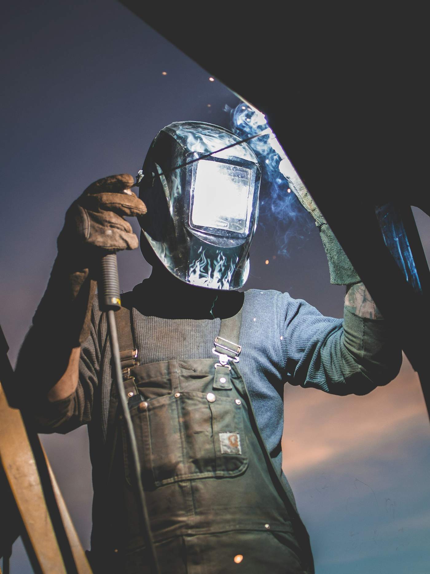 A welder in a protective mask and apron, seen against the sky